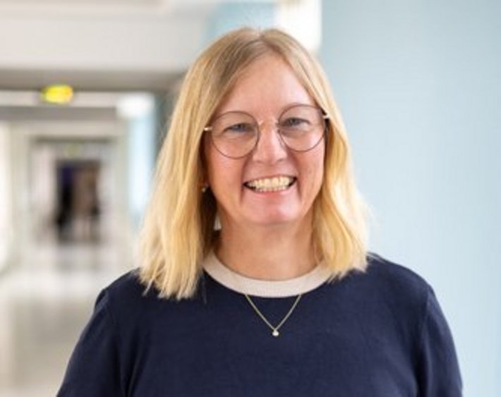 Sandra Münch (© Johannes Lehner) Blonde Dame mit mittellangen Haaren in blauem Shirt mit weißen Bündchen, Kette und Brille, lachend am Flur