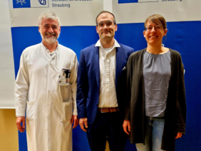 Der Chefarzt links im Arzt-Kittel mit weißem Haar und Bart neben dem Oberarzt in Jackett, dunkel und weißes Hemd, Brille und der Oberärztin mit Brille in Jeans, alle freundlich lächelnd vor zwei Rollups mit dem Logo des Klinikums