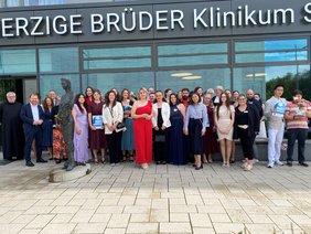 Examensfeier 2025 (© Ursula Eisenmann) Die Gruppe steht vor dem verglasten Eingang mit großem Schriftzug des Kliniknamens darüber. Viele Mädchen in Kleidern, in der Mitte eines in Rot. Der Pater Provinzial in schwarzer Ordenskutte.