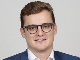 Portrait eines Herrn ohne Bart, mit Brille und kurzen braunen Haaren, blauem Sacko und weißem Hemd, lächelnd
