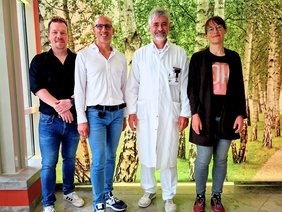 (© Alexandra Marquardt) Vor einem landschaftlich grünem LED Bild alle in Jeans, nur Prof. Jacob mit weißem Bart in weiß und Arztkittel. Alle freundlich lächelnd.